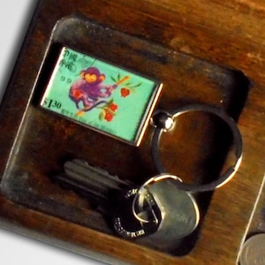 Peut inclure: Un porte-clés en bois avec un porte-clés, des clés et des pièces de monnaie. Le porte-clés a un petit charme rectangulaire coloré avec un singe dessus. Le charme a le texte "Pine Tree" et "1,30 $" dessus.