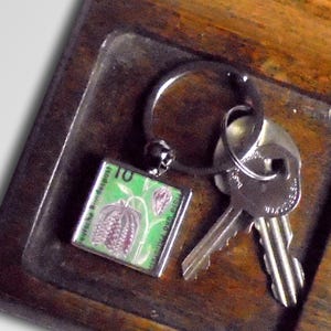 May include: Wooden key holder with a key ring, two keys, and several coins. The key ring has a small square charm with a green and purple flower design and the number 10.