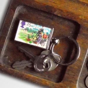 May include: A wooden tray with a key chain, keys, and several US coins. The key chain features a small rectangular charm with a colorful image and the number "25" printed on it.
