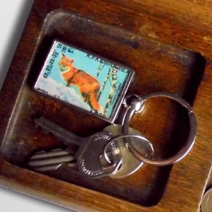 May include: A wooden tray with compartments holding keys, a keychain with a fox image, and various coins. The keychain features a rectangular image of a fox in a snowy landscape. The coins include a half-dollar and several smaller denominations.