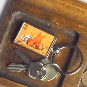 May include: A wooden tray with a key chain featuring a small metal charm with a Chinese stamp depicting a dog. The tray also has a set of keys and several US coins.