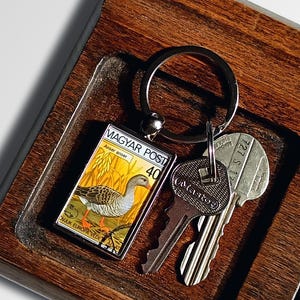May include: A wooden tray with a key chain featuring a vintage Hungarian postage stamp with a bird design. The tray also has a set of keys and several coins.