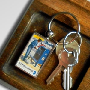 Könnte beinhalten: Ein Holztablett mit einem Schlüsselanhänger mit einer alten US-Postmarke, zwei Schlüsseln und einer Sammlung von Münzen.