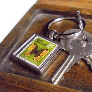 May include: A silver key chain with a small, square, green and yellow butterfly stamp charm. The charm has the text "New Zealand 2c" on it. The keychain is hanging on a wooden key holder.