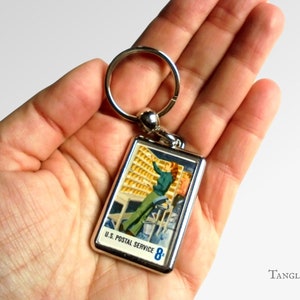 Könnte beinhalten: Ein silberner Schlüsselanhänger mit einer alten US-Postmarke, auf der ein Postangestellter beim Sortieren von Post zu sehen ist. Die Marke trägt die Aufschrift "U.S. Postal Service 8c".