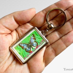 May include: A silver key chain with a green and blue butterfly on a green background. The butterfly is surrounded by a silver frame and the text "REP. OF ZAMBIA" is printed on the frame.