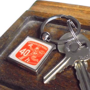 Könnte beinhalten: Ein silberner Schlüsselanhänger mit einem quadratischen Anhänger in Rot und Weiß, auf dem "Geschwindigkeitsbegrenzung 40 h" steht. Der Anhänger ist an einem silbernen Schlüsselring mit zwei silbernen Schlüsseln befestigt.