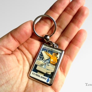 Könnte beinhalten: Ein silberner Schlüsselanhänger mit einem rechteckigen Bild eines Postbeamten, der Post sortiert. Das Bild trägt den Text "U.S. Postal Service 8¢".