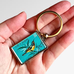 May include: Keyring with a rectangular pendant depicting a bird illustration on a teal background. The pendant has a gold-coloured frame and a gold keyring. The text "VIET NAM" is visible on the pendant.