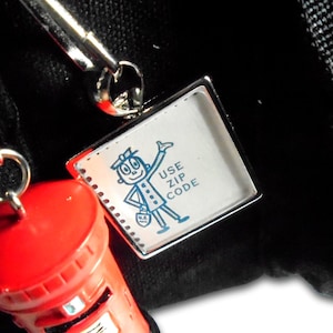 May include: A keychain featuring a red pillar box charm and a silver split ring and lanyard clip. The keychain includes a small rectangular charm with a cartoon figure and the text "USE ZIP CODE". The text "Your keychain will come with both split ring & lanyard clip" is also visible.