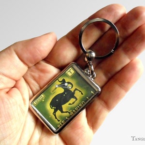 May include: A rectangular metal keychain featuring a green background with a black ram silhouette representing the Aries zodiac sign. The design includes the text ARIETE and REP S MARINO, along with a small constellation graphic and symbol.