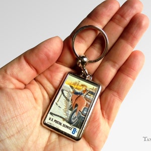 Könnte beinhalten: Ein silberner Schlüsselanhänger mit einem rechteckigen Anhänger, der eine Vintage-Illustration eines Postwagens zeigt, der durch Schnee fährt. Der Anhänger trägt den Text "U.S. Postal Service 8¢".