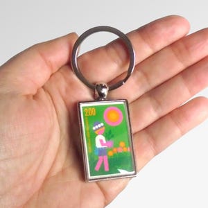 May include: A silver key chain with a green, orange and pink rectangular charm featuring a woman holding a plant and the text "200"