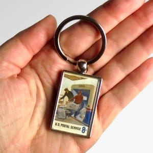 Könnte beinhalten: Ein silberner Schlüsselanhänger mit einem rechteckigen Metallanhänger, der einen Vintage-Briefmarken der US Postal Service mit einem blau-weißen Design zeigt. Die Briefmarke zeigt zwei Postangestellte, die Post in einen LKW laden. Der Anhänger trägt den Text "U.S. Postal Service 8¢".