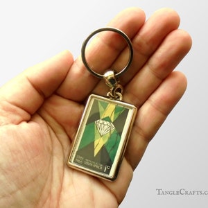 May include: A silver metal keychain with a rectangular charm featuring a green, yellow, and black diamond design. The charm has the text "1961 TRANSVAAL 1c 1966 SOUTH AFRICA" printed on it.