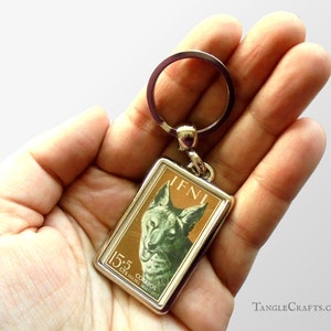 May include: A silver keychain with a rectangular metal charm featuring a vintage postage stamp design. The stamp depicts a cat with the text "15.5 Cts. INAME MALAGASY" and "IFNI" in the upper right corner.