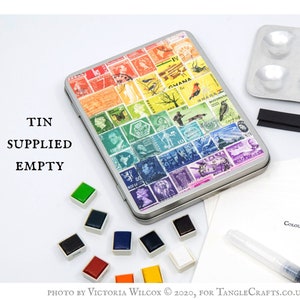 May include: An empty metal tin featuring a colourful collage of vintage postage stamps. The tin is surrounded by watercolour paint pans and a brush.