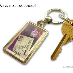 May include: A silver keyring with a rectangular charm featuring a vintage Italian postage stamp with the text "POSTE" and "CODICE AVVIAMENTO POSTALE 125". The charm is attached to a silver split ring with a gold-coloured key ring. The text "KEYS NOT INCLUDED!" is visible at the top of the image.