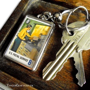 Könnte beinhalten: Ein silberner Schlüsselanhänger mit einem Vintage-Bild eines Postbeamten an einem Schalter. Das Bild ist in Silber gerahmt und trägt den Text "U.S. POSTAL SERVICE 8¢". Der Schlüsselanhänger ist an einem Schlüsselsatz befestigt.
