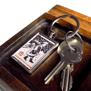 Könnte beinhalten: Ein silberner Schlüsselanhänger mit einem kleinen, rechteckigen, gerahmten Bild eines japanischen Briefmarken-Vintage mit einem Mann in rotem Mantel und Hut. Die Marke ist mit "15" und "NIPPON" beschriftet. Der Schlüsselanhänger ist an einem Holztablett mit einem Schlüssel und Münzen befestigt.