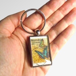May include: A silver key chain with a rectangular charm featuring a stamp from Namibia with two butterflies. The stamp says "Standardised Mail" and "Namibia".