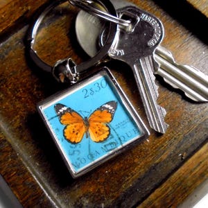 May include: A square keychain with a butterfly design. The butterfly is orange and black, set against a light blue background. The keychain is attached to a silver key ring and a set of keys. The text "2$30" is visible.