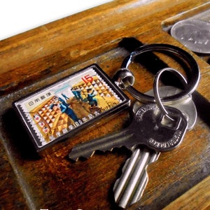 Könnte beinhalten: Ein silberner Schlüsselanhänger mit einer japanischen Briefmarke mit einem Nennwert von 15 Yen und dem Wort "NIPPON" auf einem Holztablett mit Münzen.