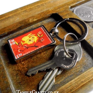 May include: A silver key ring with a red and gold Chinese New Year stamp featuring a rabbit and the text "2011 Year of the Rabbit". The keyring is on a wooden tray with UK coins.