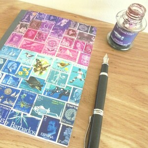 May include: A notebook with a cover made of colourful postage stamps from around the world. The notebook is lying on a wooden surface next to a black fountain pen and a bottle of ink.