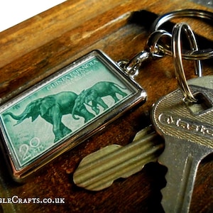 May include: A rectangular silver keychain featuring a green postage stamp design with two elephants. The stamp reads "GUINEA ESPAÑOLA" and "20 CTS". The keychain is attached to a keyring with keys.