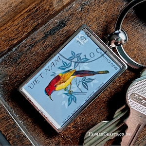 May include: A rectangular keychain featuring a colorful bird design on a light blue background, with text in Vietnamese. The keychain has a silver metal frame and a key ring attached. The bird is yellow, red, and green.
