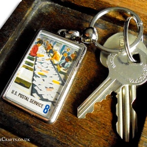 Könnte beinhalten: Ein silberner Schlüsselanhänger mit einem kleinen rechteckigen Anhänger, der ein Vintage-Bild einer Postsortiermaschine zeigt. Der Anhänger trägt den Text "U.S. Postal Service 8¢". Der Schlüsselanhänger ist an zwei silbernen Schlüsseln befestigt.