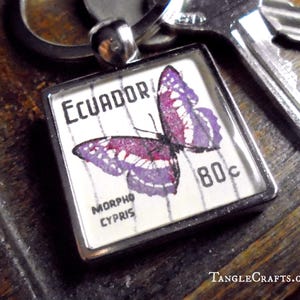 May include: A square keychain pendant featuring a vintage Ecuador postage stamp with a purple butterfly design. The stamp reads "ECUADOR" and "80c" with the text "MORHO CYPRIS". The pendant is in a silver-tone metal frame.