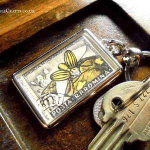 May include: A silver key chain with a clear plastic charm featuring a vintage Romanian postage stamp with a yellow and black butterfly and the text "GINDACUL DE COLORADO - LEPIDOPTERES - POSTA R.P.ROMINA".
