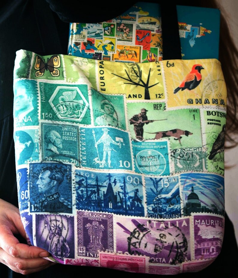 May include: A colourful tote bag with a patchwork design featuring vintage postage stamps from around the world. The bag has a green, blue, and white background with images of birds, trees, and ships. The text on the stamps includes "Ghana", "Botswana", "Helvetia", "Rep. E", "Europa", "United States Postage", "Italia", "Mauritius", "Service Postage", "India", and "United States Postage".