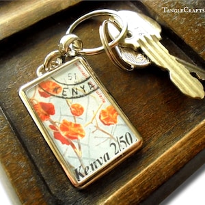 May include: A rectangular keychain with a gold-colored frame, featuring a vintage Kenya stamp with orange floral imagery. The keychain is attached to a silver keyring with two keys resting on a wooden tray.