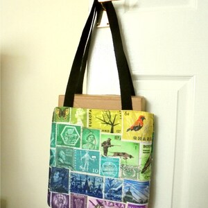 May include: A black tote bag with a colourful design featuring postage stamps from around the world. The bag has a rainbow gradient with a variety of stamps, including a red bird, a tree, and a ship. The text "EUROPA" and "ITALIA" are visible on the stamps.