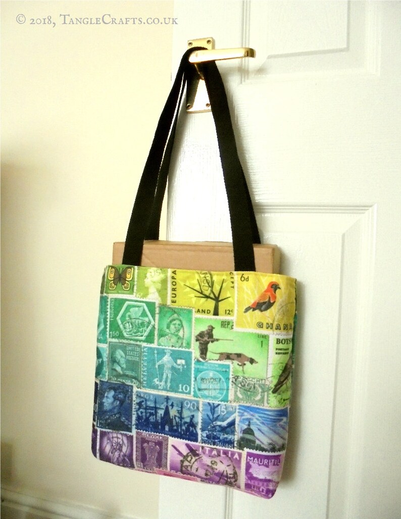 May include: A black tote bag with a colourful design featuring postage stamps from around the world. The bag has a rainbow gradient with a variety of stamps, including a red bird, a tree, and a ship. The text "EUROPA" and "ITALIA" are visible on the stamps.