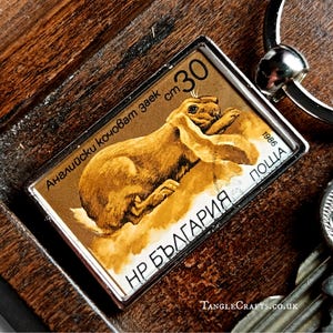 May include: A rectangular keychain featuring a vintage postage stamp design with a brown rabbit illustration. The stamp reads "30 cm" and "1986". The keychain has a silver-colored metal frame and a key ring.