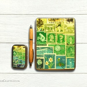 May include: Two rectangular metal tins featuring a collage of vintage postage stamps in shades of green, yellow, and blue. The larger tin is 18 cm tall and the smaller tin is 10 cm tall. A wooden pen is lying next to the tins.