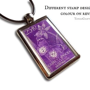 May include: A purple stamp pendant necklace featuring a butterfly and flower design. The stamp reads "GUINEA ESPAÑOLA CORREOS DIA DEL SELLO 1958" and "25-10 CTS".