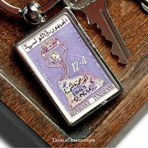 May include: A rectangular keychain with a silver frame featuring a vintage Tunisian postage stamp design. The stamp is predominantly purple with Arabic text and a stylized figure. The keychain is next to a key on a wooden surface.