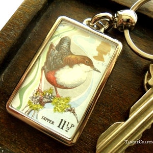 May include: A silver key chain with a rectangular charm featuring a vintage illustration of a dipper bird on a branch. The charm has a blue and white background with the text "DIPPER 1 1/2 P" printed on the bottom.
