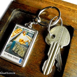 Könnte beinhalten: Ein silberner Schlüsselanhänger mit einem kleinen rechteckigen Anhänger, der eine Vintage-Illustration einer Postsortiermaschine zeigt. Der Anhänger trägt den Text "U.S. Postal Service 8".