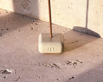 May include: A square, off-white incense holder with a carved design, holding a brown incense stick. The holder is made of a solid material and sits on a textured surface. The incense stick is upright.
