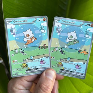 Op de afbeelding: Twee Cubone EX Pok&eacute;mon-verzamelkaarten. Elke kaart toont een cartoon Cubone-personage met een oranje of groene outfit, tegen een achtergrond van blauwe lucht en groen gras. De kaarten hebben 70 HP.