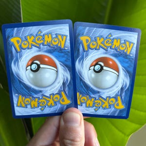 Op de afbeelding: Twee Pok&eacute;mon-verzamelkaarten met blauwe achterkanten, met het Pok&eacute;mon-logo in geel en een Pok&eacute; Ball-ontwerp. De kaarten worden tegen een achtergrond van groene bladeren gehouden.