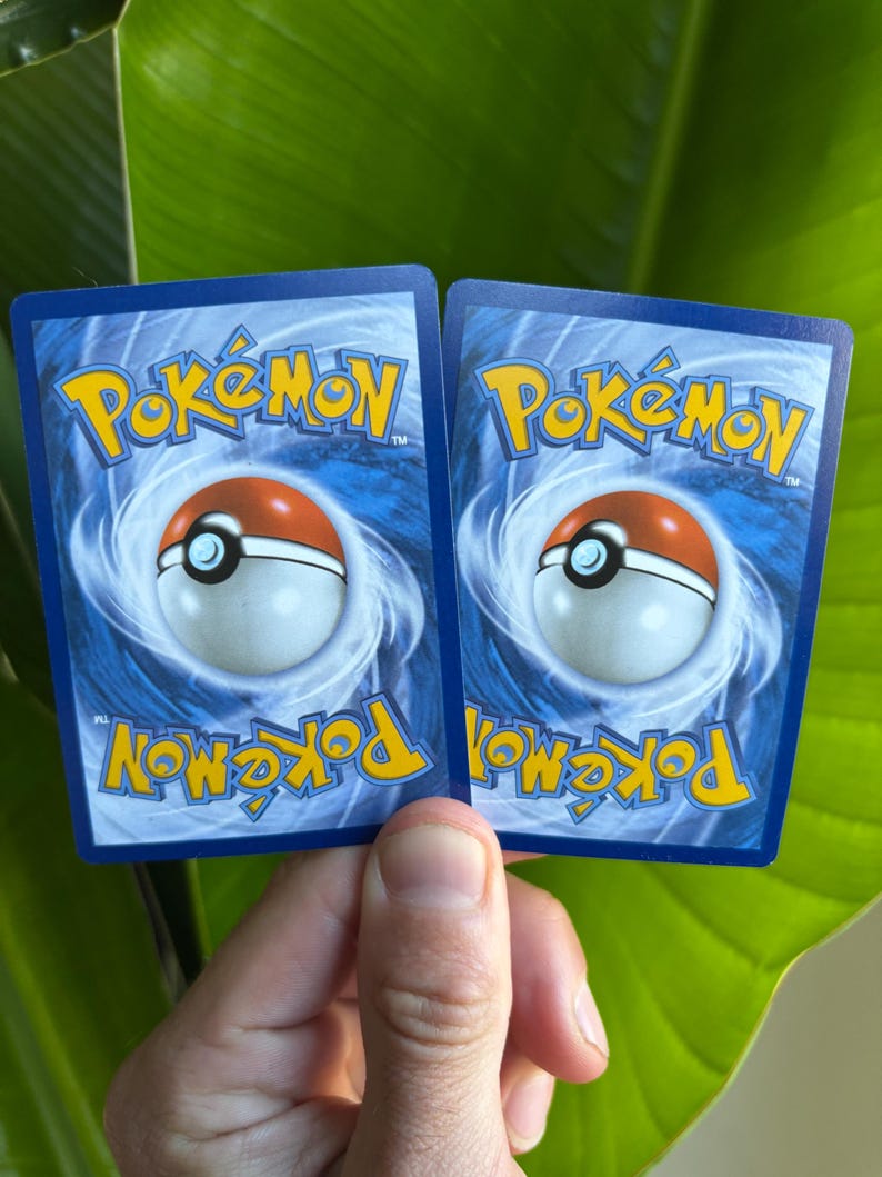 Op de afbeelding: Twee Pok&eacute;mon-verzamelkaarten met blauwe achterkanten, met het Pok&eacute;mon-logo in geel en een Pok&eacute; Ball-ontwerp. De kaarten worden tegen een achtergrond van groene bladeren gehouden.
