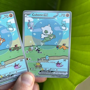 Op de afbeelding: Twee Cubone EX Pok&eacute;mon ruilkaarten. De kaarten tonen een wit Cubone-personage met een groene en blauwe achtergrond. De kaarten hebben de tekst "Cubone EX", "Beat" en "Bone Smash".