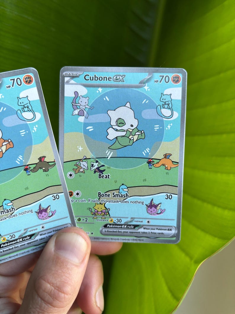Op de afbeelding: Twee Cubone EX Pok&eacute;mon ruilkaarten. De kaarten tonen een wit Cubone-personage met een groene en blauwe achtergrond. De kaarten hebben de tekst "Cubone EX", "Beat" en "Bone Smash".